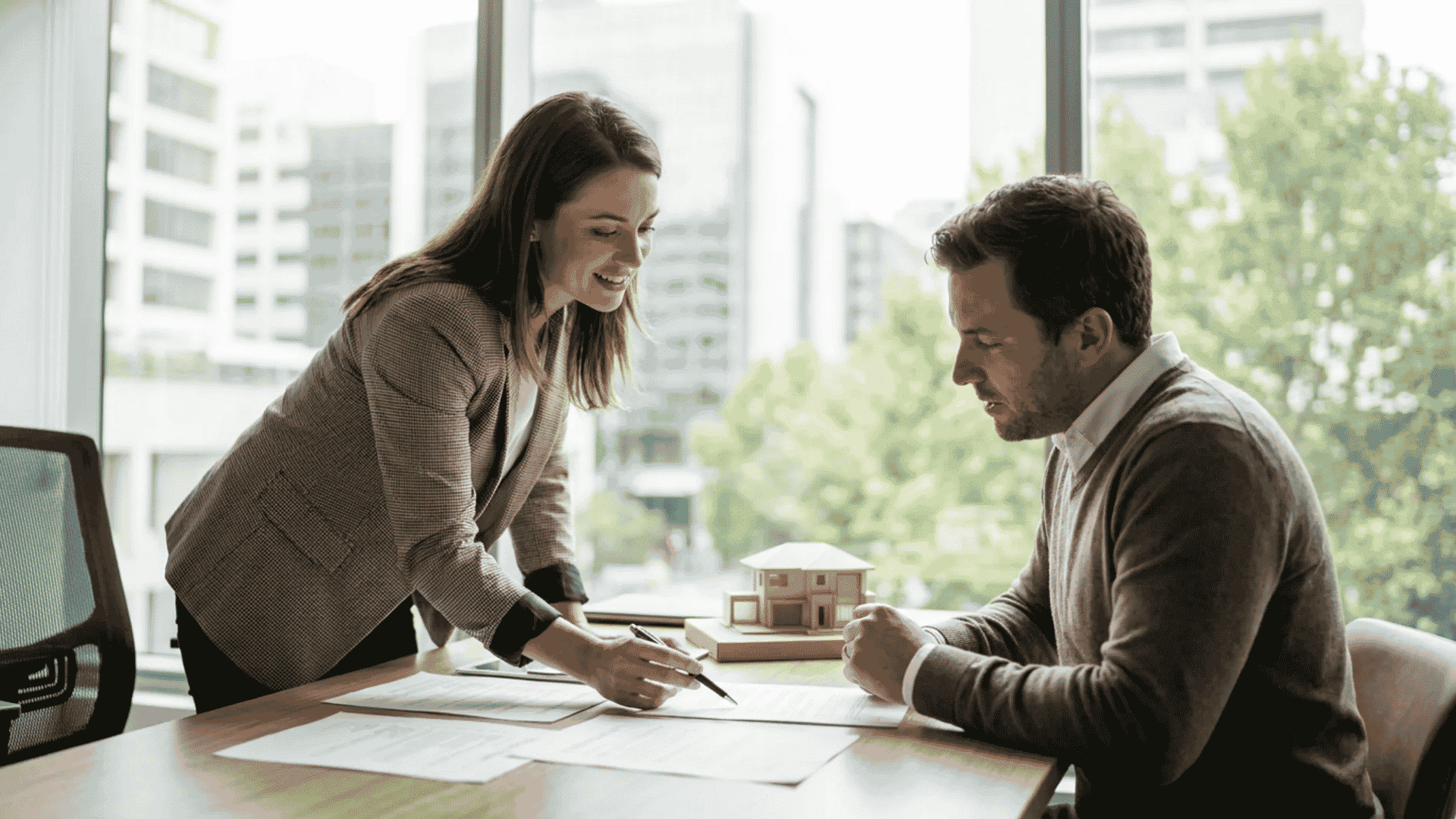 A mortgage advisor discussing paperwork and a tiny house model with a client at an office desk near a window.