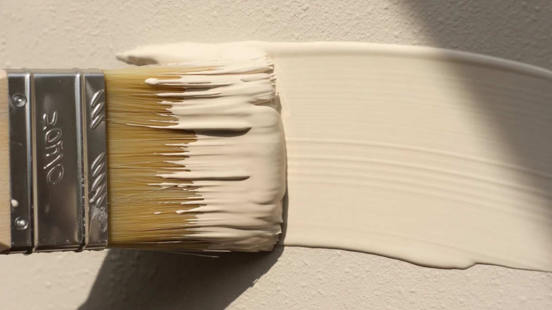 Close-up of a paintbrush applying smooth beige water-based paint evenly across a textured wall surface.