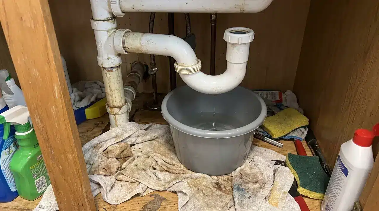 White PVC P-trap pipe being removed under a kitchen sink with a bucket underneath.