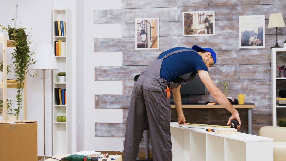 Carpenter measuring and assembling furniture for a media wall installation in a modern living room.