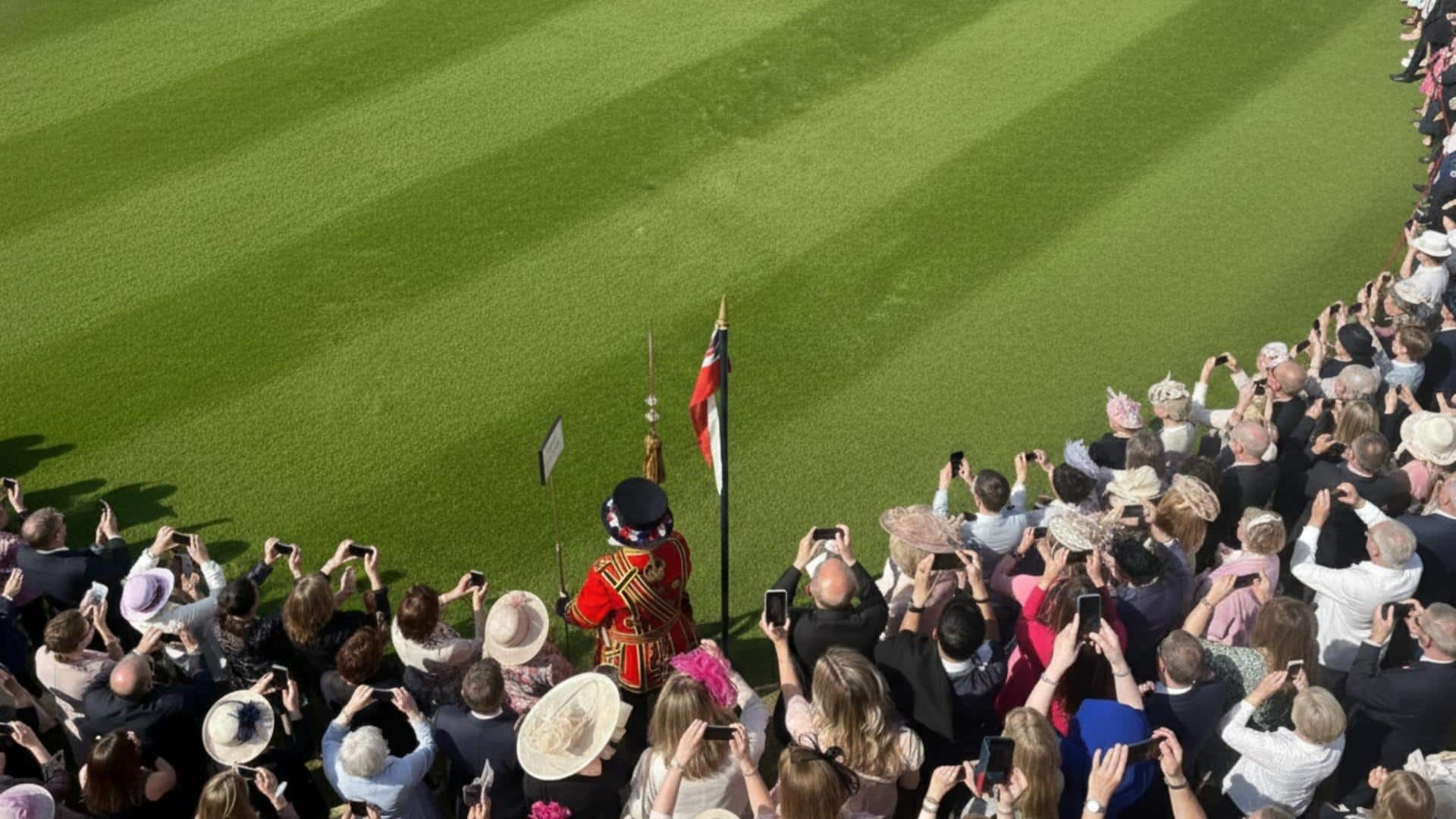A ceremonial guard in red uniform standing on a lawn before a crowd of formally dressed guests.