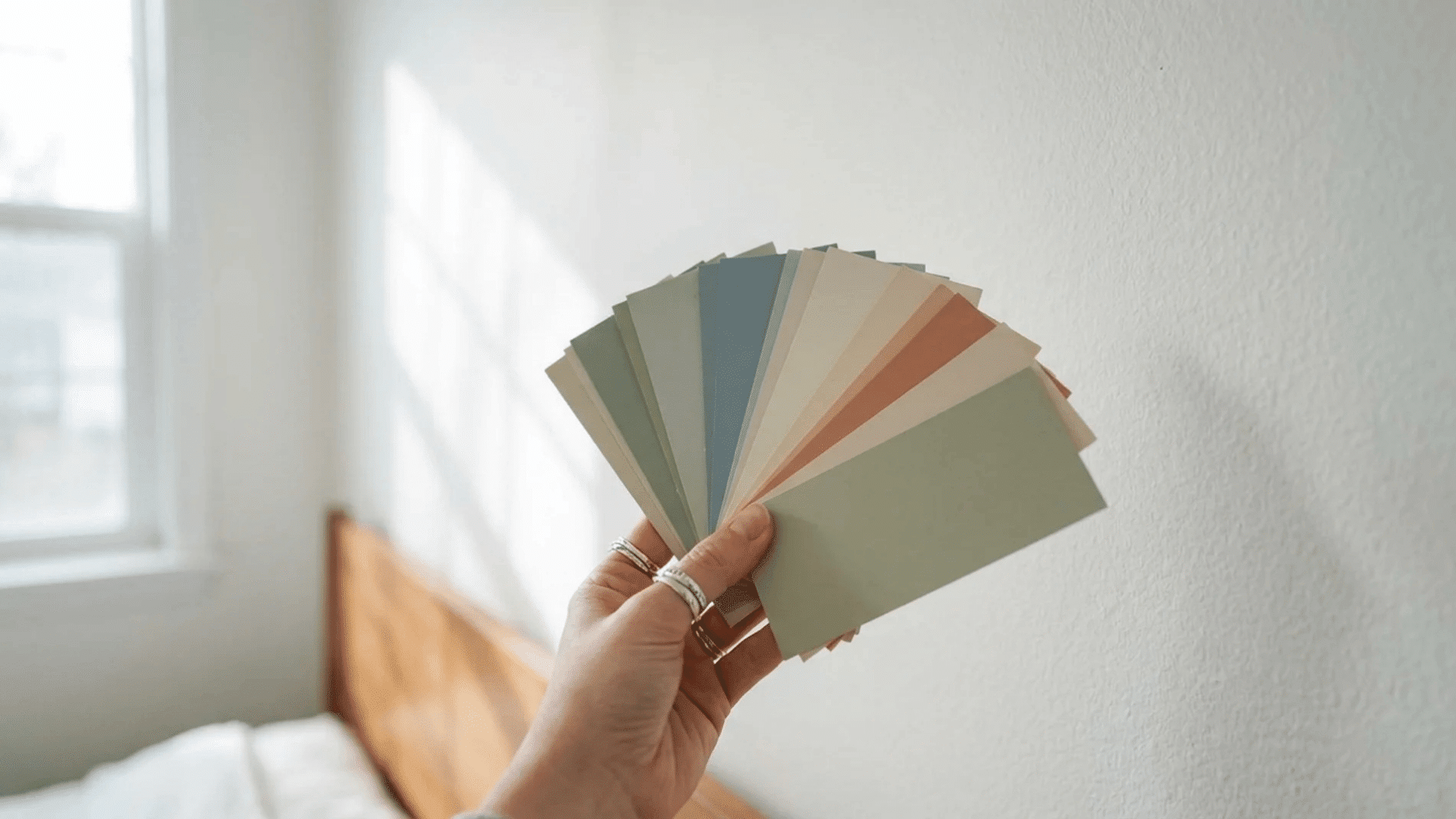 Hand comparing paint color swatches against a bedroom wall under natural light.