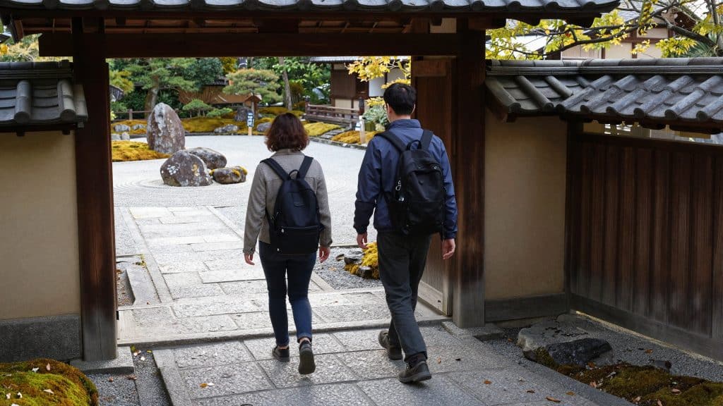Visitors walking through a traditional Japanese garden entrance toward a peaceful Zen rock garden with autumn foliage
