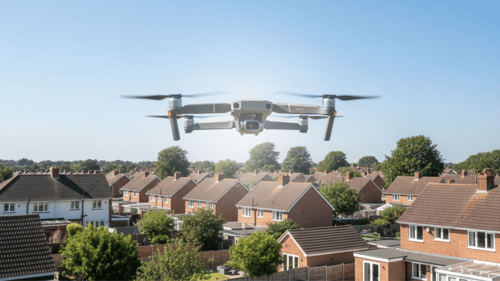 Drone flying above a suburban UK neighborhood with rows of brick houses beneath a clear blue summer sky