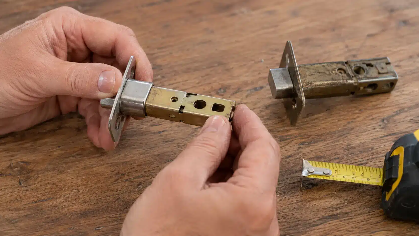 Hands adjusting a new extendable door latch beside an old latch and tape measure on a wooden workbench.