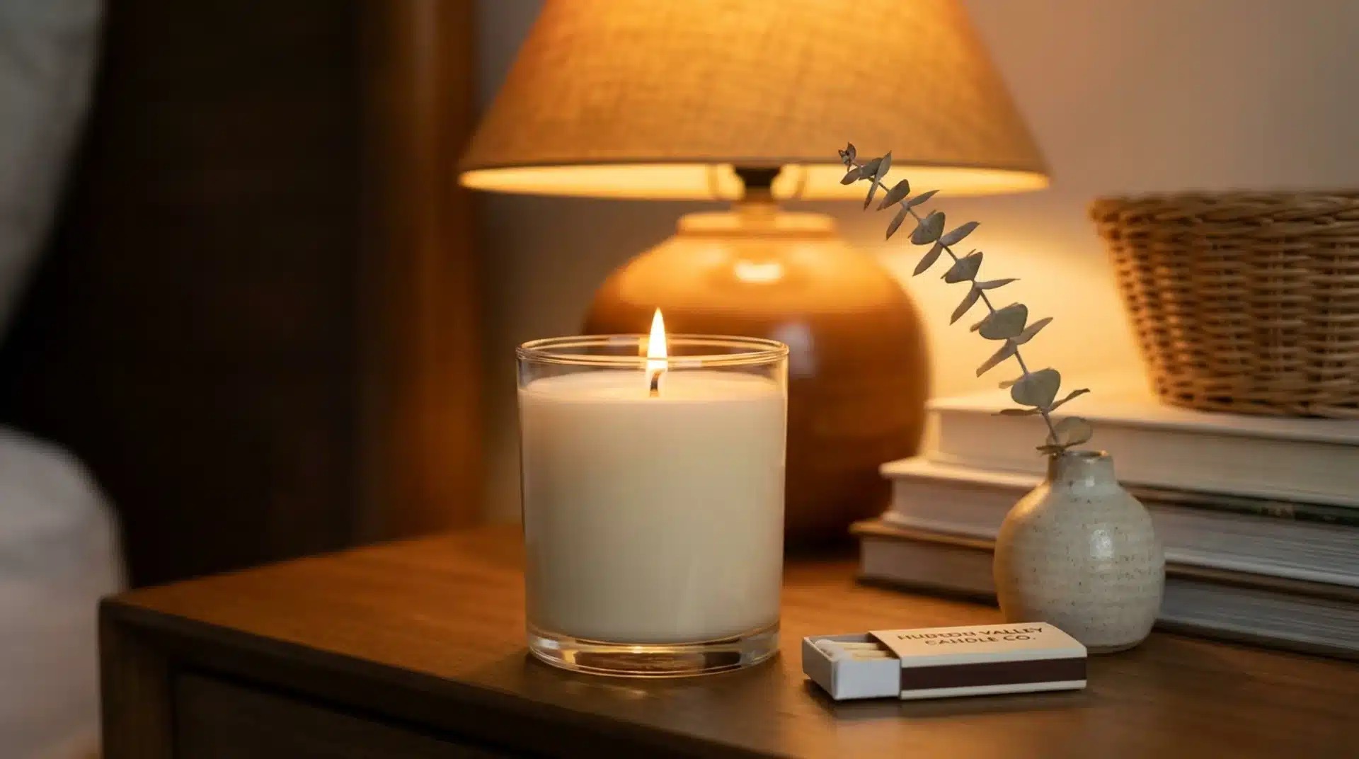 Guest bedroom nightstand with a cream candle, matchbox, and dried stem bud vase in warm amber light.